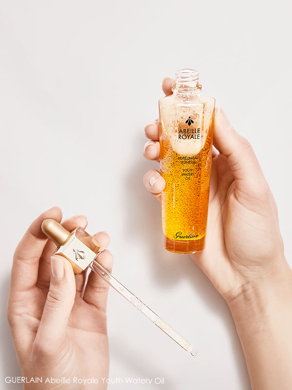 Two hands holding an open bottle of the GUERLAIN Abeille Royale Youth Watery Oil